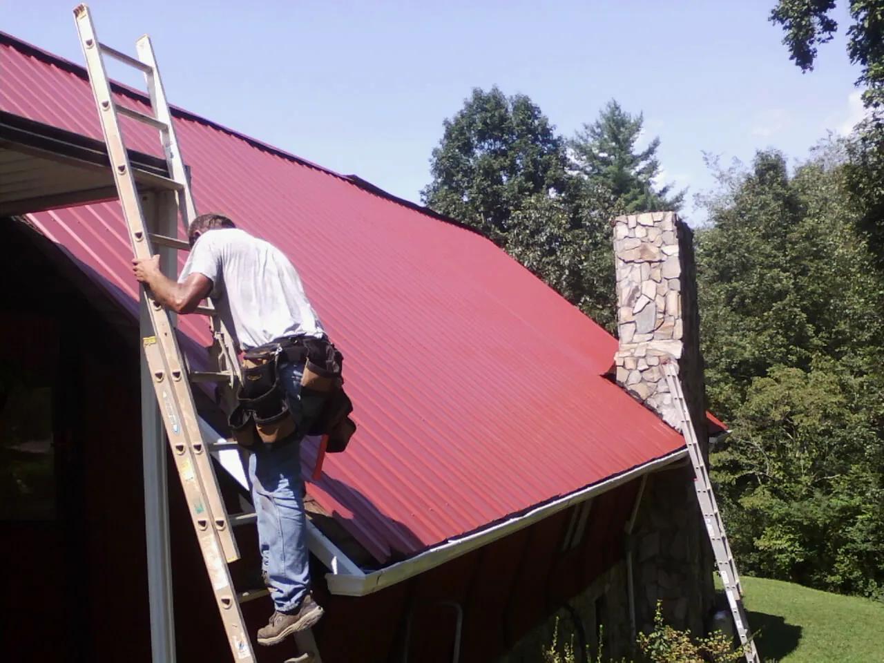 Roofer on a ladder beside a red metal roof and stone chimney — Soffit & Fascia Repair by Roofing Brothers in Crested Butte